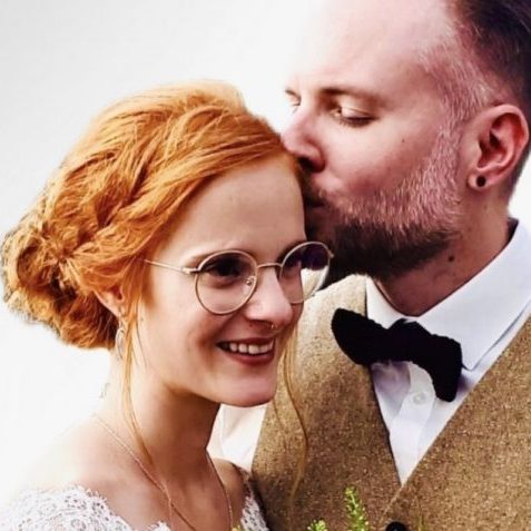 Brautpaar: die Frau mit rotem Haar und Brille, der Mann küsst sie liebevoll auf die Stirn.