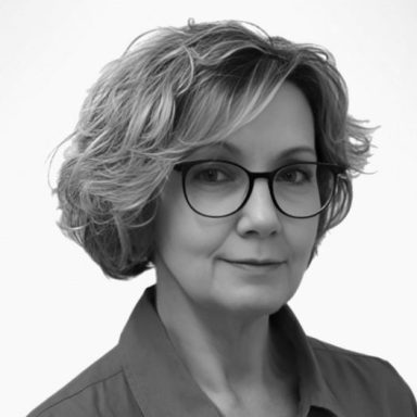 Stefanie Kamin Porträt einer Frau mit lockigem Haar und Brille, in neutralem Hintergrund.